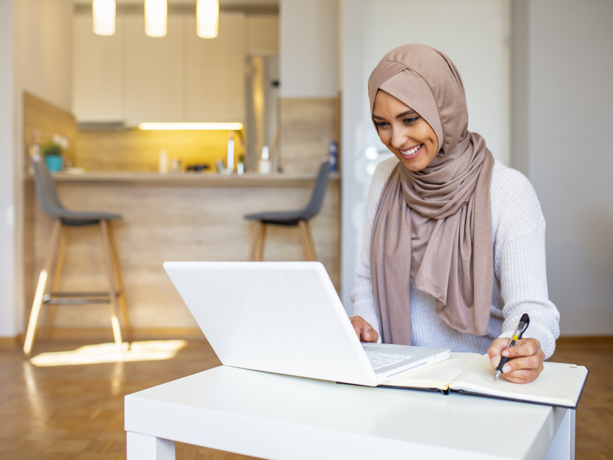 woman wearing a head scarf working from a laptop and taking notes